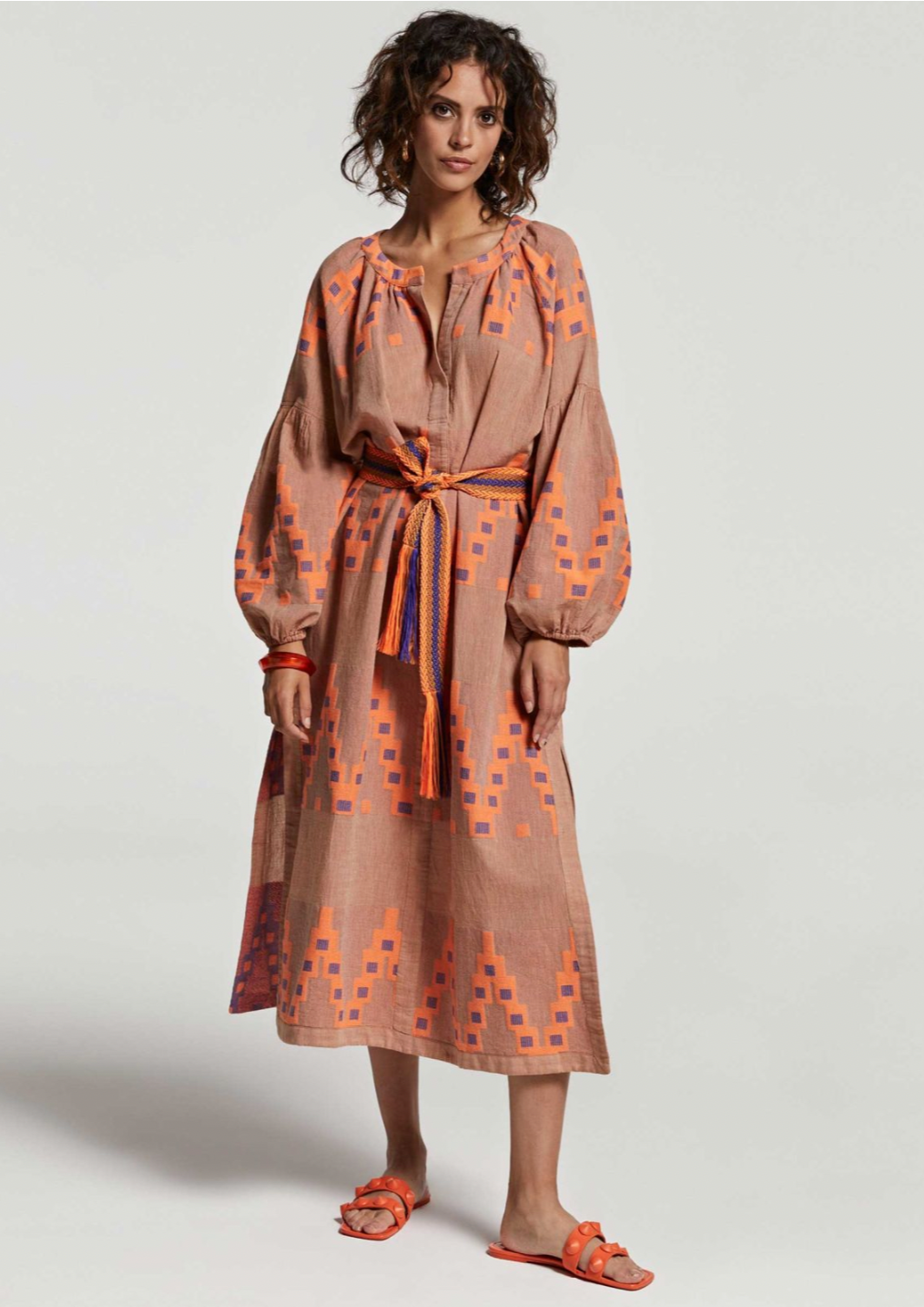 Devotion Korali Dress - Beige/Orange The Korali Maxi dress from Devotion Twins is a vacation essential. Made with an airy cotton blend and featuring Devotion's distinct geometric jacquard pattern all over, this piece is complete with a snap button detail V-neck and a woven waist belt. Dropped shoulder balloon sleeves and side slits add the finishing touch to this relaxed look.  Wear free and easy, or belt to elevate your look for evenings out